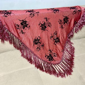 May include: A pink shawl featuring black floral designs and a shimmering fringe. The shawl is displayed over a neutral surface, highlighting its triangular form and decorative details. The fringe provides texture and visual interest to the piece.