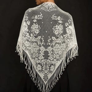 Puede incluir: Un chal triangular de encaje blanco con un intrincado patrón floral sobre un fondo de malla fina. Los bordes del chal están rematados con largos y delicados flecos que cuelgan de la punta inferior y de los lados de la prenda.