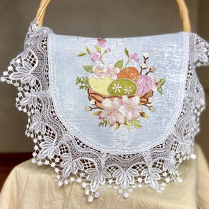 May include: A light blue Easter basket liner with a floral and egg embroidery design. The embroidered design features a basket of colourful Easter eggs and pink flowers. The liner is trimmed with white lace.