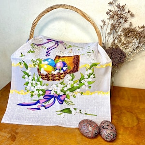 May include: A wicker basket with a handle contains a light grey Easter-themed cloth. The cloth depicts a basket of decorated eggs, a cake, and lily of the valley flowers. Two decorative Easter eggs with intricate patterns are on a wooden surface.