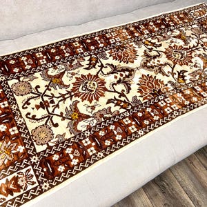 May include: Rectangular rug with a cream-colored center featuring floral and botanical patterns in shades of brown and gold. The border is a darker brown with geometric designs. The rug is placed on a light gray surface.