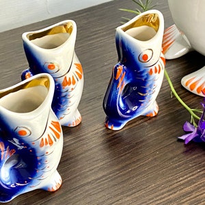 May include: Three blue and white porcelain fish-shaped shot glasses with orange accents. The glasses are arranged on a wooden surface.