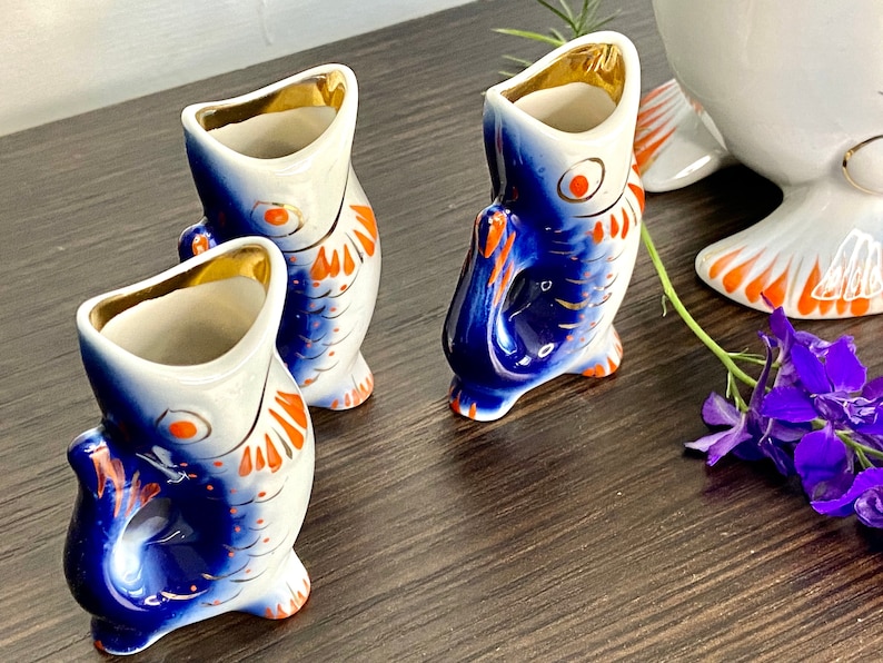 May include: Three blue and white porcelain fish-shaped shot glasses with orange accents. The glasses are arranged on a wooden surface.