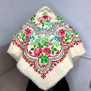 May include: A square, cream-coloured scarf with a floral design. It features large pink roses, green leaves, and small blue flowers, framed by a red border. The scarf has a fringed edge.