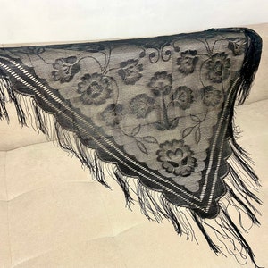Può includere: Scialle in pizzo nero con ricamo floreale e bordatura a frange. Lo scialle è drappeggiato su una superficie di colore chiaro.