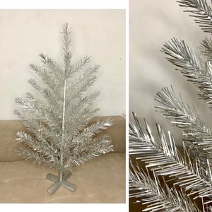 Puede incluir: Un árbol de Navidad de aluminio plateado con una base en forma de cruz. El árbol tiene un diseño moderno clásico de mediados de siglo, con ramas metálicas que se extienden desde el poste central. Una decoración festiva.