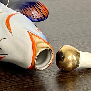 May include: A white porcelain fish bottle stopper with gold and orange accents. The stopper is in two pieces, the top is a round gold piece and the bottom is a white piece.