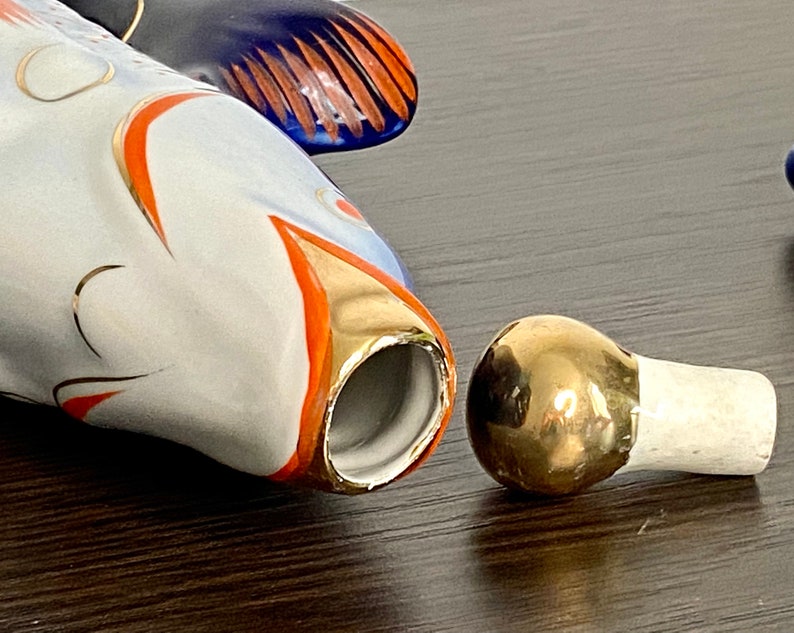 May include: A white porcelain fish bottle stopper with gold and orange accents. The stopper is in two pieces, the top is a round gold piece and the bottom is a white piece.