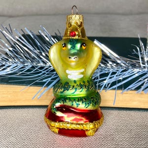 Könnte beinhalten: Ein gläserner Weihnachtsbaumschmuck in Form einer grünen und gelben Kobra mit einer roten Krone und auf einem roten Kissen sitzend. Der Schmuck ist mit goldenen Verzierungen verziert.