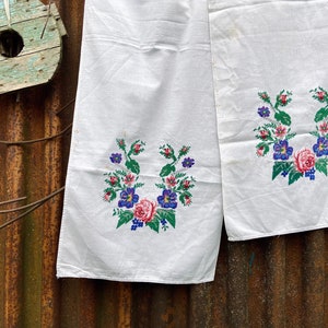 May include: Two white linen towels with embroidered floral designs. The embroidery features pink roses, blue and purple flowers, and green leaves. The towels are hanging against a weathered, corrugated metal background.