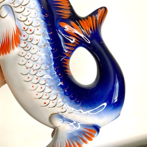 May include: A blue and white porcelain fish-shaped pitcher with orange and gold accents. The pitcher has a detailed design of scales and fins.