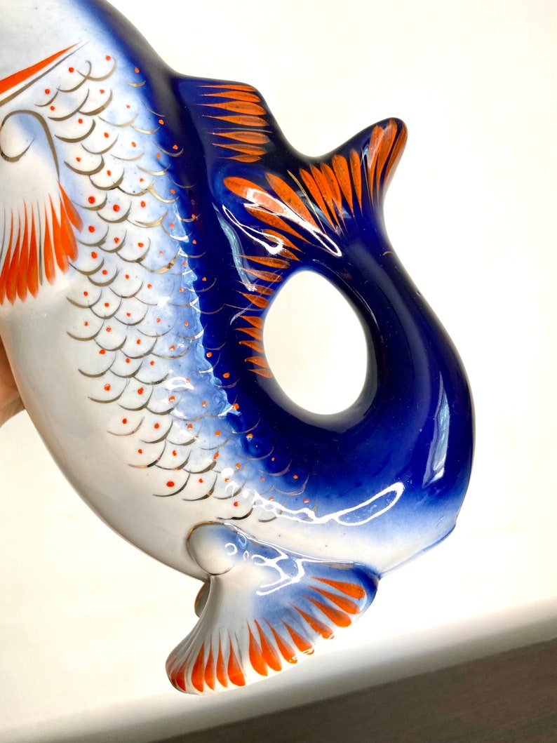 May include: A blue and white porcelain fish-shaped pitcher with orange and gold accents. The pitcher has a detailed design of scales and fins.