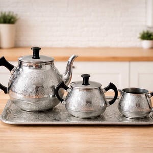 Könnte beinhalten: Ein silbernes Teeservice mit Teekanne, Zuckerdose und Sahnekännchen auf einem passenden Tablett. Das Set hat schwarze Griffe und Deckel sowie ein geprägtes Blumendesign. Das Set wird auf einer Holzoberfläche präsentiert.