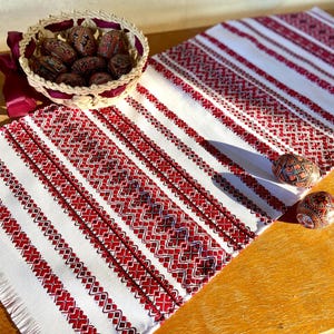 May include: A white table runner with red embroidered stripes and a fringed border. The runner is decorated with a traditional Ukrainian pattern. Two red and gold painted Easter eggs are on a wooden table next to the runner.