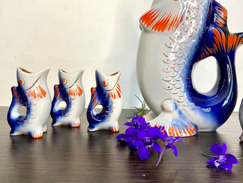 May include: Set of four blue and white porcelain fish-shaped shot glasses with orange and gold accents. The set includes a larger fish-shaped pitcher with a blue and white design.