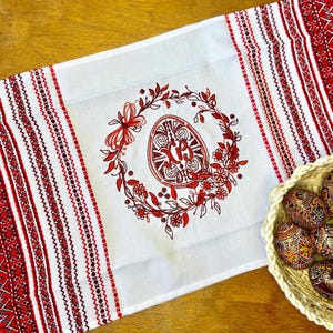 May include: White Easter table runner with red embroidered floral and geometric patterns. A central design features a red egg within a wreath. A basket of decorated Easter eggs sits to the right.