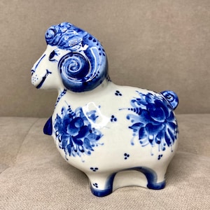 May include: A ceramic ram figurine featuring a white base with hand-painted blue floral patterns and intricate swirling details on the horns. The ram has a glossy finish, a small tail, and blue accents on the hooves, face, and neck area.