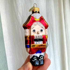 May include: A decorative glass Christmas ornament shaped like a nutcracker soldier. The ornament features a red and gold uniform, a white face with a sad expression, and black boots. The ornament has a gold metal cap and a loop for hanging.
