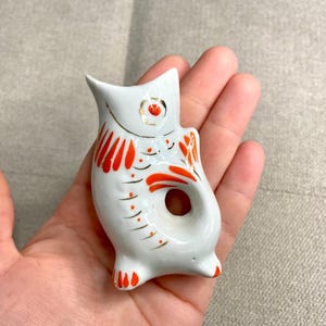 May include: White ceramic fish figurine with red and gold painted details. The fish has a hole in the middle, suggesting it may be a bottle stopper.