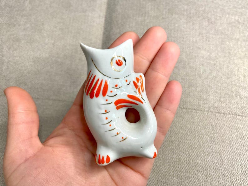 May include: White ceramic fish figurine with red and gold painted details. The fish has a hole in the middle, suggesting it may be a bottle stopper.