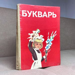 May include: A vintage book with a red cover featuring the Cyrillic word "БУКВАРЬ" in white. The cover illustration depicts a young girl in a school uniform holding a bell and flowers. The book's spine is off-white.