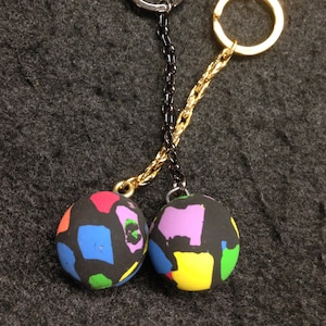 May include: Two keychains with black, round pendants featuring colorful abstract designs. Each pendant is attached to a chain and a metal ring. One keychain has a silver ring and black chain, the other has a gold ring and chain.