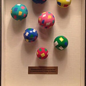 May include: A framed display of eight colorful balls with a diamond pattern. The balls are arranged in a circle and are displayed on a white background.