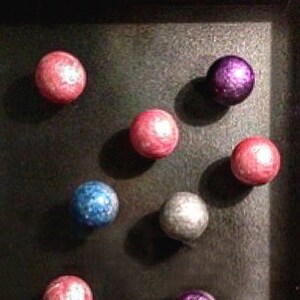 Può includere: Una teca con cornice nera che contiene nove sfere di vetro colorate. Le sfere sono disposte a formare un motivo e sono di varie tonalità di rosa, viola, blu e argento.