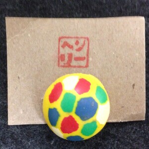 May include: A small, round, yellow ball with red, blue, green, and white spots. The ball is sitting on a brown paper card with a red stamp that says "ペー" in Japanese.