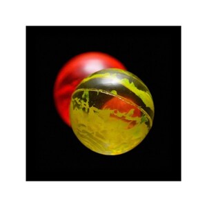 May include: A yellow rubber ball with red and green paint splatters. The ball is in focus and a red ball is out of focus in the background.