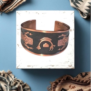 May include: A copper cuff bracelet with a dark gray center band featuring embossed designs. The bracelet has a wide, open design and is displayed on a white surface against a blue background. Woven textiles with geometric patterns are in the corners.