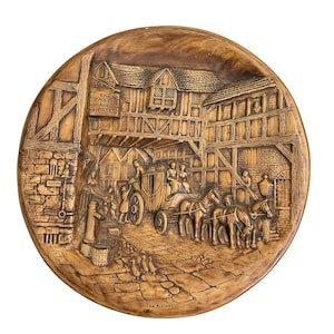 May include: A round brown wall plaque featuring a relief scene of a horse-drawn carriage arriving at an inn. The sign reads The Coach and Horses, and the bottom text reads The Arrival. The scene includes stone walls, timber framing, and horses.