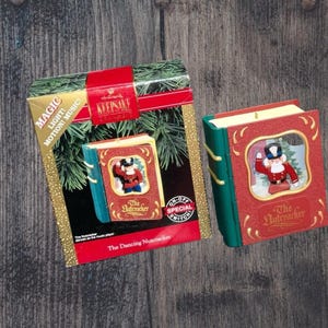 May include: A Hallmark Keepsake ornament in the shape of a book, featuring a red cover with a framed image of a Nutcracker soldier. The ornament has an on/off switch and is packaged in a box with text that reads "The Dancing Nutcracker".