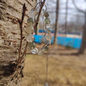 May include: A silver wire bracelet with clear and green glass beads. The bracelet is hanging from a tree branch.