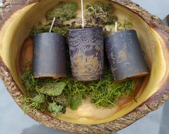 May include: Three dark gray pillar candles with white wicks are nestled in a wooden bowl filled with green moss. The bowl has a natural, rustic edge and a light yellow interior. The candles have a textured appearance.