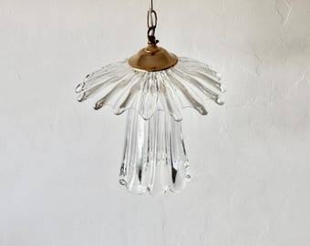 Pode incluir: Luminária pendente de vidro transparente com abajur franzido e fixação cor de latão. A luminária pende de uma corrente e é projetada com um corpo canelado em forma de tulipa. A luz é suspensa contra uma parede branca.
