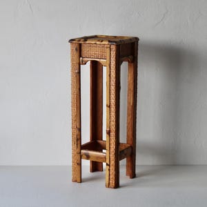 May include: A tall, rectangular plant stand made of woven rattan and wood. The stand has a square top with a woven surface and a lower shelf. The legs are sturdy and the stand is a natural brown colour.