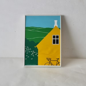 May include: Framed art print depicting a yellow house with a white chimney and a black window. A black bicycle is in front of the house. The background shows green hills and a blue sky. The artwork has a minimalist style.