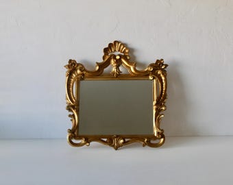 Pode incluir: Um espelho retangular dourado com uma moldura ornamentada em estilo barroco. A moldura apresenta detalhes intrincados de volutas e florais, com uma crista decorativa no topo. O espelho está sobre um fundo branco.