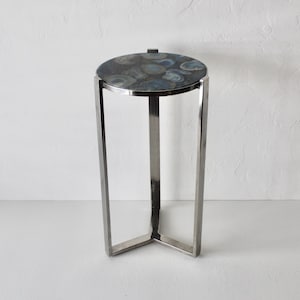 May include: A modern side table with a round top and a stainless steel frame. The tabletop features a dark, patterned surface with shades of blue and gray. The table stands approximately 24 inches tall.