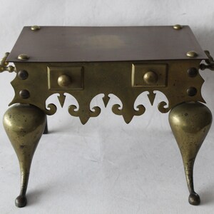 May include: A brass fireplace stand with a square top and three legs. The stand has a decorative design around the bottom edge and a knob on each side.