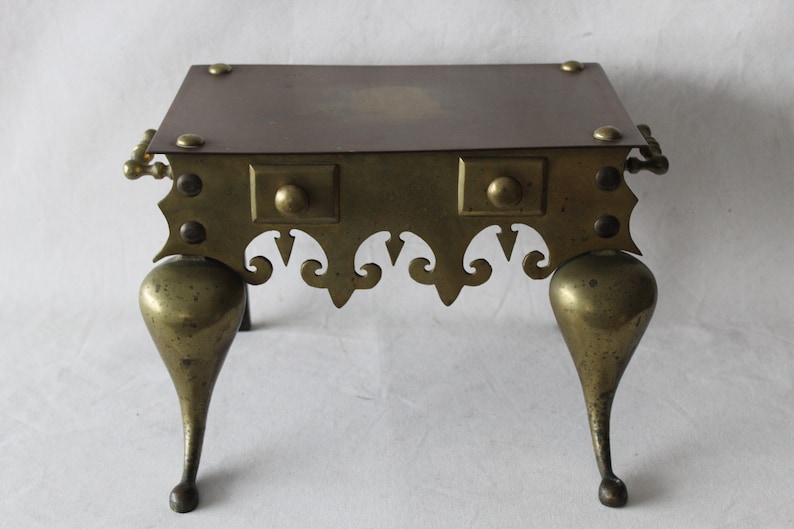 May include: A brass fireplace stand with a square top and three legs. The stand has a decorative design around the bottom edge and a knob on each side.