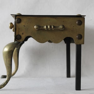 May include: A brass and iron stand with a scalloped edge and a decorative handle. The stand has three brass legs and two iron legs.
