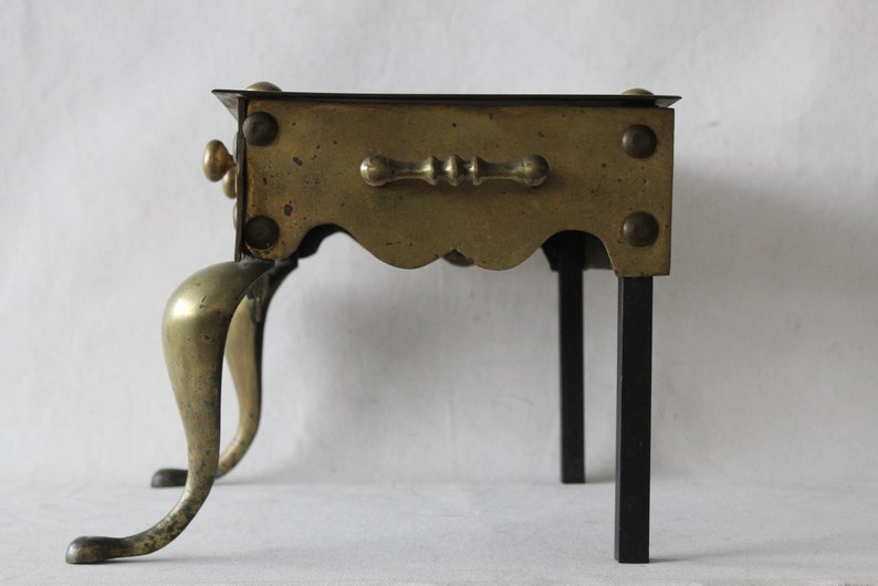 May include: A brass and iron stand with a scalloped edge and a decorative handle. The stand has three brass legs and two iron legs.