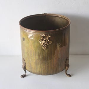 May include: An antique brass-toned metal container with a rounded body and three decorative feet. The container has a detailed gold-colored emblem on the front and a dark interior. The exterior has a mottled green and brown finish.