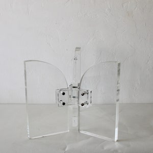 Puede incluir: Un sujetalibros de acrílico transparente con un diseño curvo. El sujetalibros está hecho de dos piezas que están conectadas por una bisagra. La bisagra es de metal y tiene un acabado plateado.