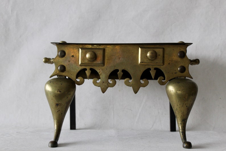 May include: A brass fireplace stand with three legs and a decorative top. The stand has a black metal frame and ornate details.