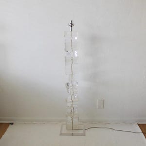 May include: A tall, clear acrylic floor lamp with a stacked design. The lamp has a simple, modern style and is made of many clear acrylic blocks.