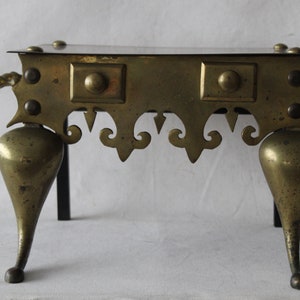 May include: A brass fireplace stand with a square top and three legs. The legs are shaped like a teardrop and have a decorative design. The stand is made of brass and has a dark patina.