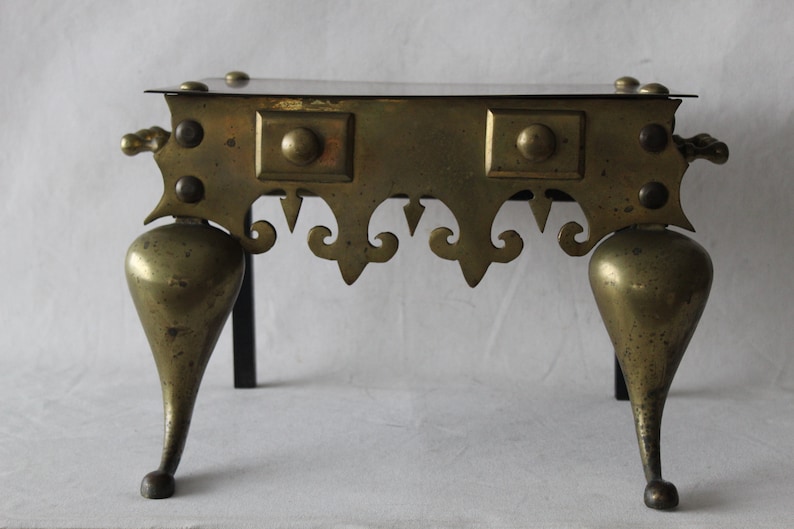 May include: A brass fireplace stand with a square top and three legs. The legs are shaped like a teardrop and have a decorative design. The stand is made of brass and has a dark patina.